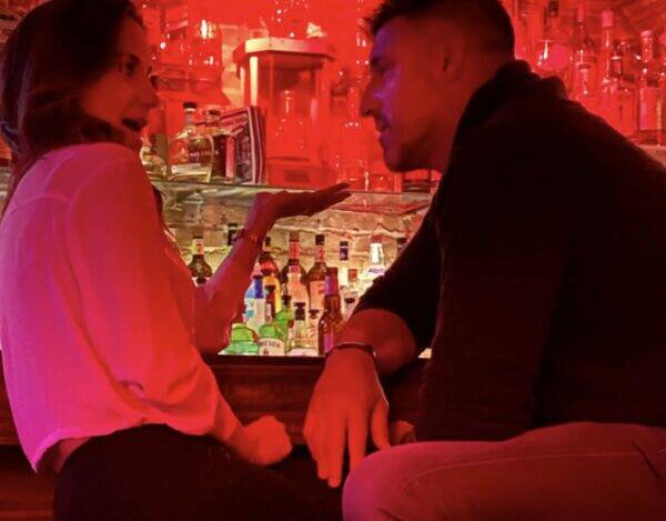 Two people sit at a bar counter, facing each other and talking, with red neon lighting and bottles lined up behind them at the bar.