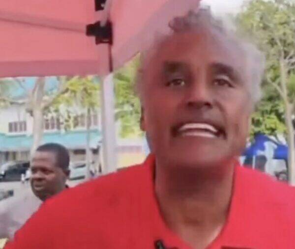 Close-up of a man with gray hair wearing a red shirt, speaking directly to the camera outdoors.