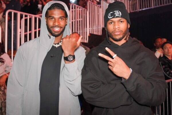 Two men posing at a crowded night event; one in a gray hooded jacket with a necklace and watch, the other in a black hoodie and beanie flashing a peace sign.