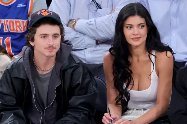 Man in a black jacket and cap sits beside a woman in a white tank top at a sports arena, both watching something off-camera.