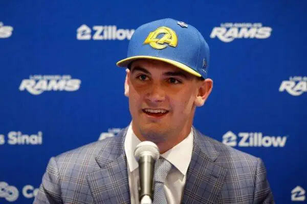 Man in a blue plaid suit speaking at a press event, wearing a blue cap with a yellow LA logo in front of a blue backdrop with sponsor logos