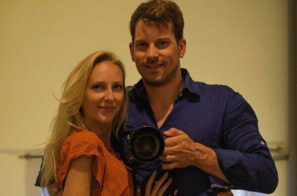 Smiling couple posing for a mirror selfie; the man holds a camera between them on a beige wall. (informative)