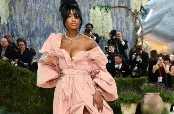 Rihanna wearing a pale pink off-the-shoulder gown with voluminous sleeves on a red carpet, posing with one hand on her hip as photographers look on.
