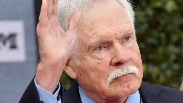 Close-up of an elderly man with a white mustache raising his right hand in a wave outdoors.