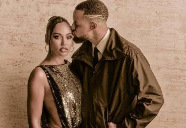 Couple posing closely; man kisses woman's temple while she looks at the camera, wearing a gold sequin dress and large hoops against a beige textured wall