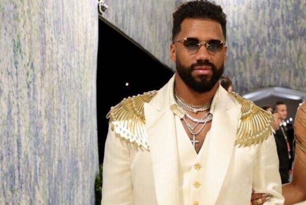 Man wearing a cream suit with gold epaulettes, multiple necklaces, and sunglasses at a formal event