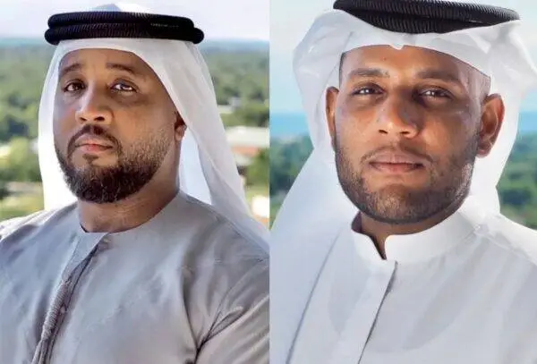 Two men wearing traditional Arab dress pose outdoors, one left with gray thawb and white ghutra, one right with white thawb and white ghutra, against a blurred background.