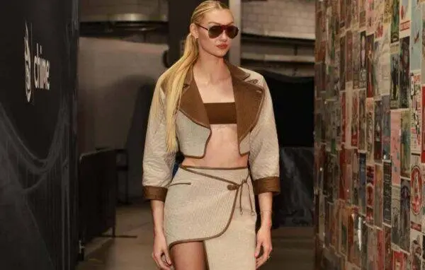 Model walking backstage in a beige and brown two-piece outfit with a cropped jacket and wrap skirt, wearing sunglasses.