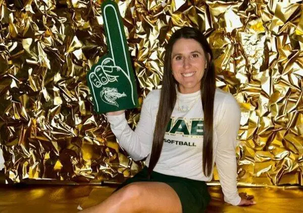 Smiling woman in a white long-sleeve shirt and green skirt sits against a gold foil background, holding a large green foam finger pointing upward.