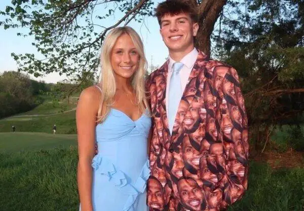 Smiling couple posing outdoors: woman in a light blue dress and man wearing a face‑print suit jacket, standing among trees and grass.