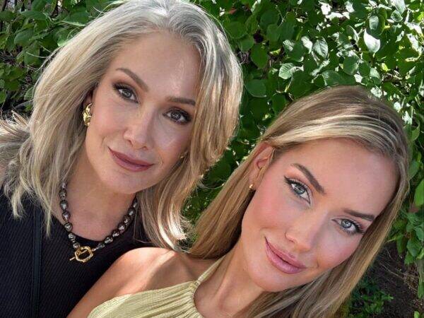 Two women posing for a close-up selfie outdoors with green foliage in the background, smiling softly.