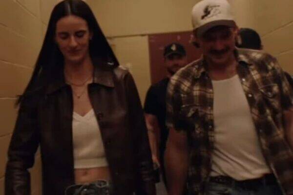 Woman in a brown leather jacket and white crop top walking beside a man in a plaid shirt and white tee down a hallway, both smiling slightly.