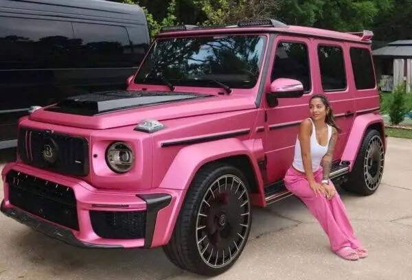 Bright pink Mercedes G-Wagon parked on a driveway, with a woman in a white tank top and pink pants sitting beside it