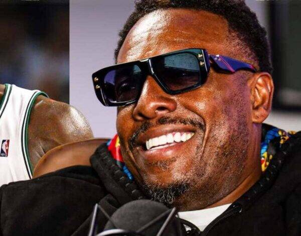 Close-up of a man smiling, wearing oversized black sunglasses, with a microphone in front.