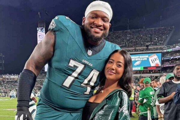 Tall football player in teal Eagles jersey #74 posing with a smiling woman on the sideline of a crowded stadium.