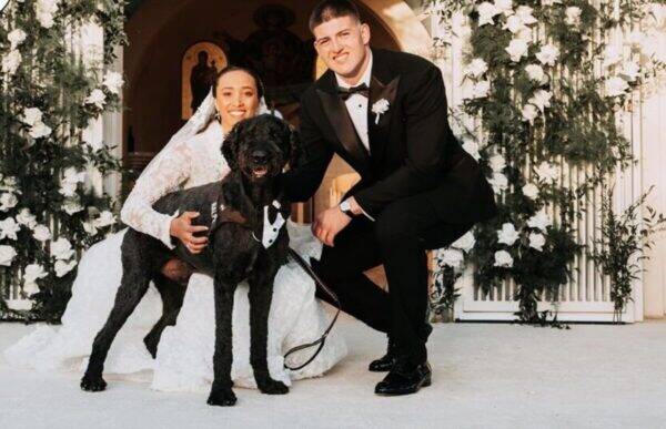 Bride and groom kneel with a black dog in a tuxedo, posed at a white floral arch.