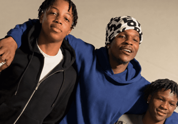 Three friends pose with arms around each other, smiling at the camera; casual hoodies and a patterned headwrap present they stand against a beige backdrop.