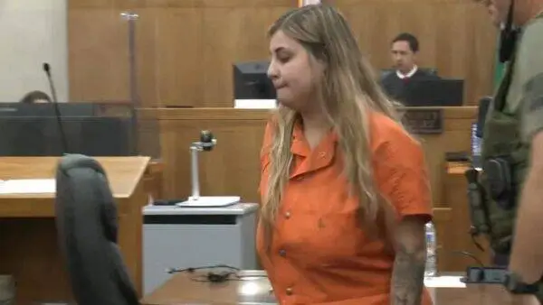 Defendant in an orange jumpsuit walks through a courtroom; a judge sits at the bench in the background.