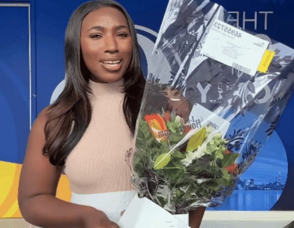 Smiling woman in a beige sleeveless top holds a large wrapped bouquet of colorful flowers in front of a blue backdrop.