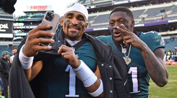 Two football players in teal jerseys take a postgame selfie on the field, one flashing a peace sign toward the camera while smiling.