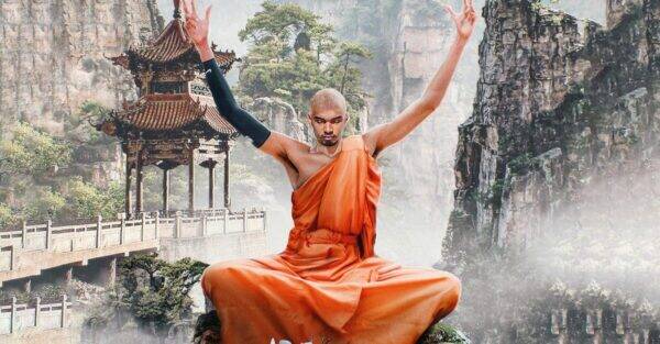 Monk in an orange robe sits cross‑legged on a rocky ledge, arms raised in meditation, with a misty canyon and pagoda behind him.