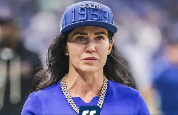 Woman wearing a blue cap embroidered with '1953' and a matching blue top, with a chunky silver chain necklace, looking off to the side.