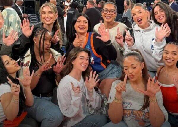 Group of diverse women posing for a group photo at a basketball event, many showing hand signs and thumbs up.