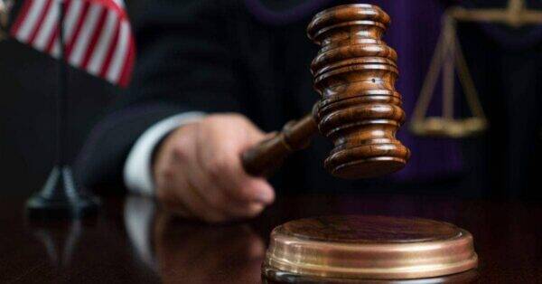 Judicial gavel raised over a sound block in a courtroom, with the scales of justice and an American flag in the background.
