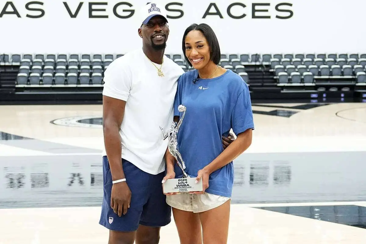 A man and a woman pose on a basketball court, holding a trophy with empty arena seating in the background.