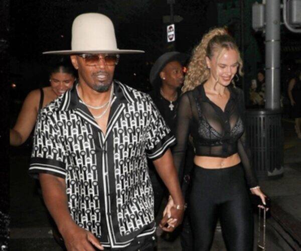 Man in a wide white hat and patterned black‑and‑white shirt walks hand‑in‑hand with a woman in a sheer black top and black leggings at night.