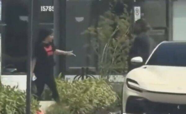 Person wearing a mask and black outfit with a pink top points toward a white car parked on the sidewalk in front of a storefront.