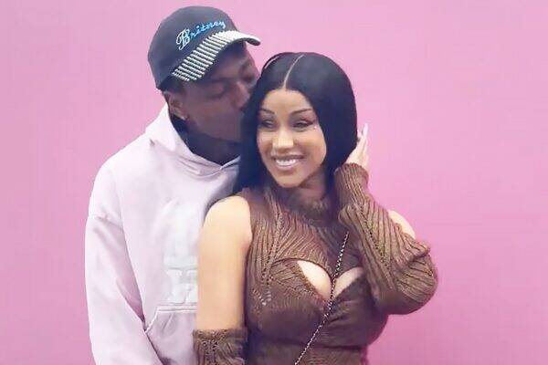 A couple stands against a pink wall; man kisses the woman's temple as she smiles at the camera.