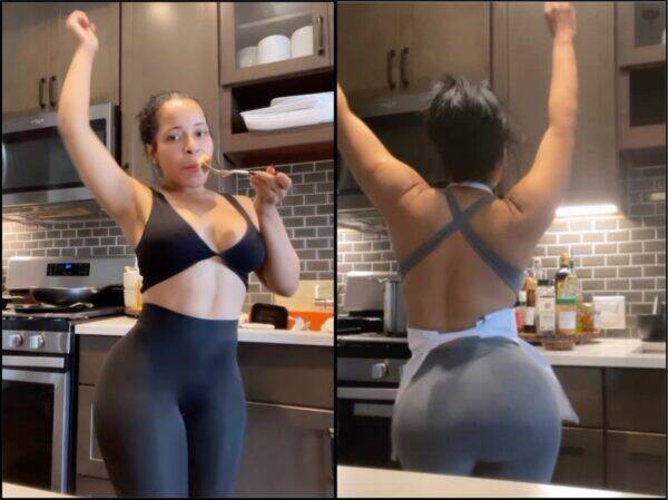 Woman in a black sports bra and leggings raises one arm while eating with a fork in a modern kitchen.