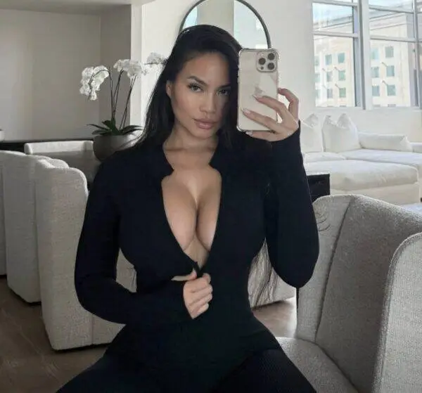 Woman with long dark hair wearing a black plunging top sits on a light sofa, taking a mirror selfie in a bright living room.