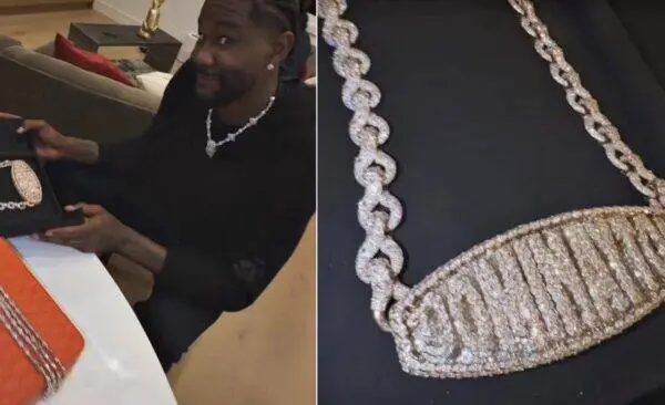 Smiling man in a black outfit shows off a sparkling diamond necklace while sitting at a table with a gift box nearby.
