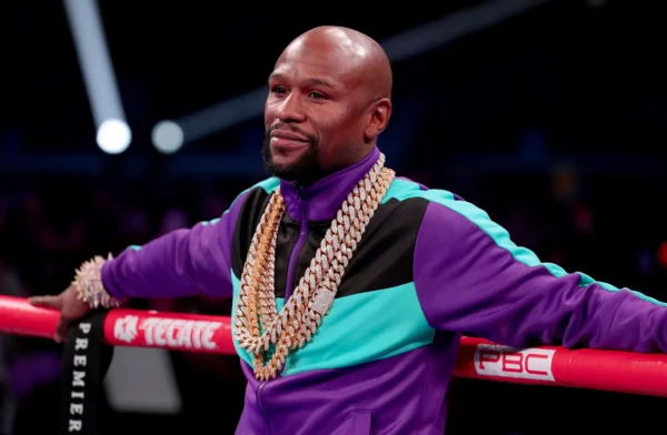 Bald man in a purple and teal tracksuit leaning on boxing ring ropes, wearing multiple chunky gold chains in a crowded arena.
