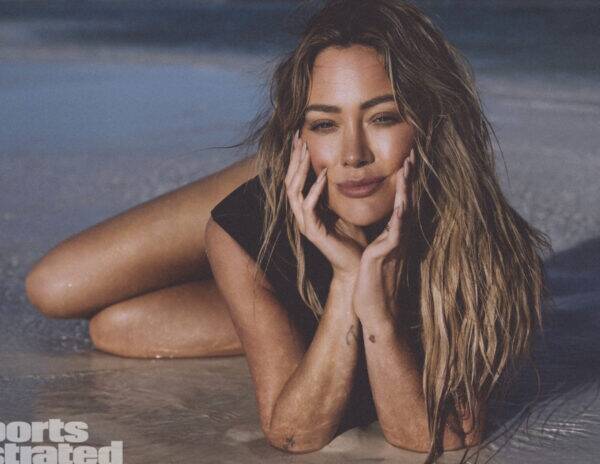 Woman with long wavy hair lying in shallow surf, hands cupping her face, wearing a black swimsuit on a sunny beach.