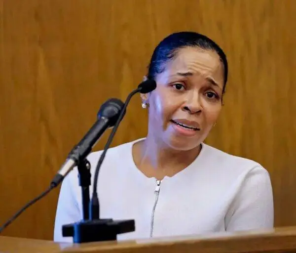 Woman in a white top standing at a wooden lectern, speaking into a microphone with a concerned expression.