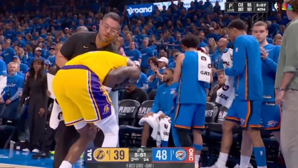 A Lakers player in a yellow uniform bends over near the sideline while a staff member leans over him; several OKC blue-clad teammates stand nearby during a timeout with a crowd in the blue seats behind them.