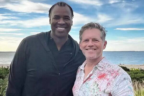 Two men smiling for a selfie on a beach, one in a black shirt and the other in a light patterned shirt, ocean and blue sky behind them.