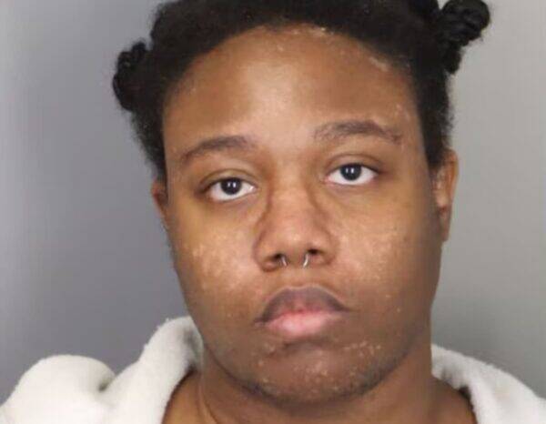Mugshot-style portrait of a young person facing the camera, wearing a light robe, with a septum piercing.