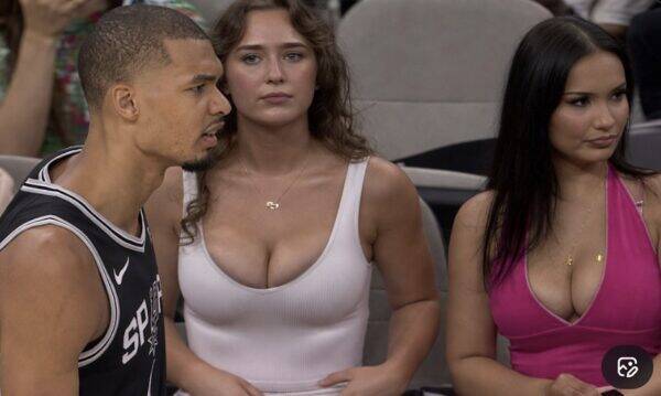 Three adults seated courtside: a man in a Spurs jersey on the left and two women in tank tops in the middle and right, looking ahead at an event.