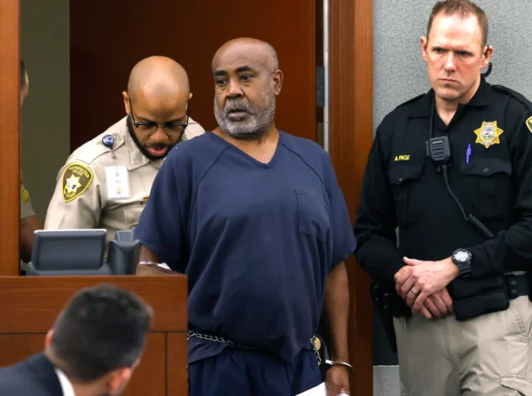 Man in blue jail uniform with handcuffs and waist chain stands in a courtroom as two uniformed officers flank him.