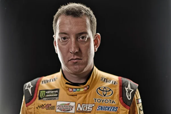 Portrait of a male race car driver looking at the camera, wearing a yellow sponsorship racing suit with logos (Toyota, Monster, Snickers) against a dark background.