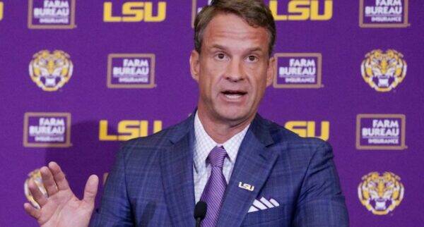 Man in a blue checked suit and purple tie speaks at a podium with an LSU backdrop behind him.