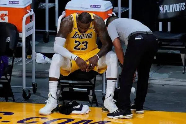 Basketball player in a Lakers yellow uniform sits on a bench, trainer leaning in as they inspect the ankle area on the floor.