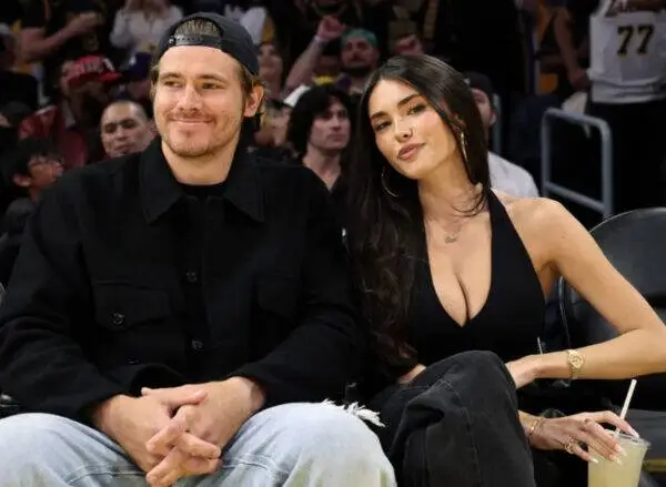 A man wearing a black jacket and cap sits beside a woman in a black sleeveless top at a sports arena, with a crowd in the background.",
