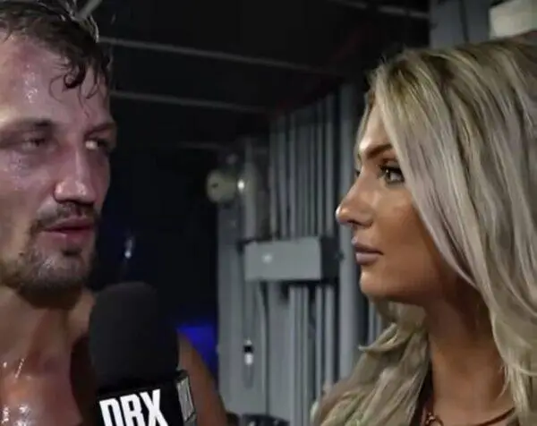 Male wrestler, sweaty and shirtless, speaks into a DRX-branded microphone during a backstage interview with a blonde female interviewer present.