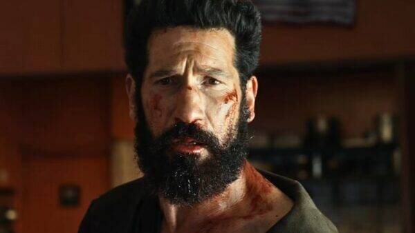 Bearded man with blood on his face and neck, looking distressed in a dim indoor setting.