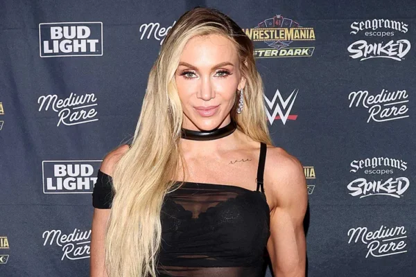 Portrait of a blonde woman posing on a red carpet at a WWE event, wearing a black sheer top and choker, with sponsor logos in the background.